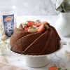 bundt cake con fragole e pistacchi su alzatina bianca accanto a barattolo di pistacchi in guscio ventura e vaso bianco sopra una tovaglia ricamata