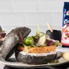 tavolo con piano in marmo su cui è posato un piatto di ceramica con panino nero con burger di salmone, salsa yogurt e insalatina, accanto a confezione chiusa di cranberry ventura e, sullo sfondo, piastrelle bianche rettangolari vintage