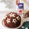 su un tavolo bianco, piatto di ceramica con panettone ricoperto di namelaka, con cranberry ventura, accanto a mani di donna con maglione bianco che reggono confezione chiusa cranberry ventura e barattolo crema alla nocciola