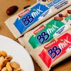 tre barrette bbmix ventura incartate azzurra verde e rossa accanto a una ciotola con frutta secca sgusciata