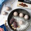 su piano di marmo, piatto scuro con tartufini dolci, farina di cocco, datteri e mandorle sgusciate e pelate, accanto a scatola di datteri medjoul ventura
