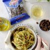 su un tavolo bianco, mano di donna curata con maglione azzurro afferra forchetta per mangiare perciati molisani pasta simile a spaghetti conditi con mollica di pane, prezzemolo e uvetta, accanto a bicchiere di vino bianco, ciotolina con uvetta, bottiglietta per l'olio e confezione chiusa di uvetta ventura