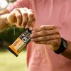 mani di uomo sportivo al parco con maglietta e smartwatch aprono confezione barretta proteica con frutta secca e arachidi