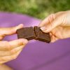 mano di uomo e mano di donna con belle unghie si dividono una barretta proteica ventura con frutta secca e cioccolato, sullo sfondo tappetino da yoga sull'erba