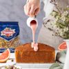mano di donna versa crema al pompelmo rosa su plumcake fatto in casa, pistacchi sgusciati ventura in busta,vaso bianco con fiori di campo