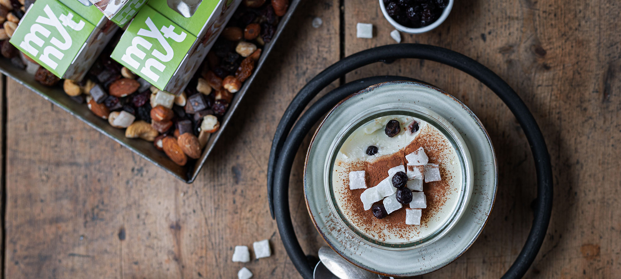 budino al latte in coppetta monoporzione con cacao amro mirtilli essiccati cocco a cubetti accanto a scatole verdi confrutta secca su tavolo di legno