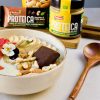 ciotola bianca con matcha bowl allo yogurt, fiori e frutta secca proteica ventura e crema spalmabile proteica alla mandorla, accanto a cucchiaio in legno