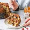 fetta di panettone con crema di nocciole e barattolo di crema gourmet ventura con nocciole con mano di donna che regge una forchetta su tavolot bianco