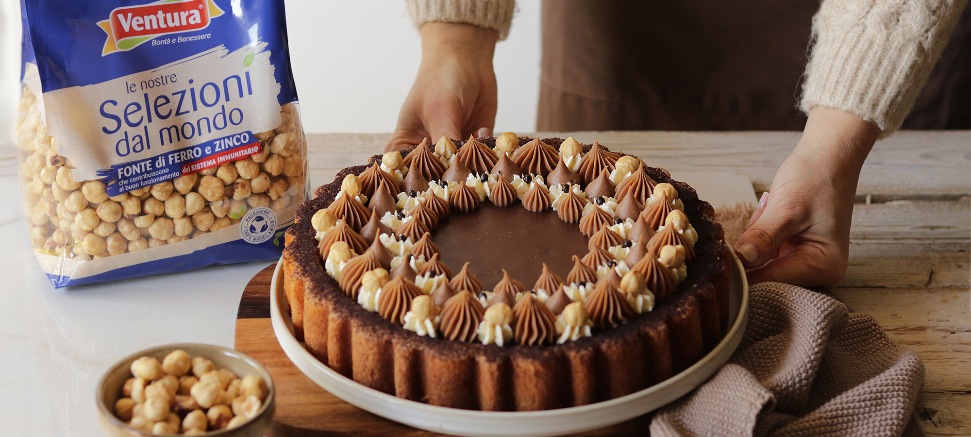 bellissima crostata morbida alle nocciole con ganache al cioccolato e decorazione bicolore su piatto da portata bianco retto da mani di donna accanto a busta di nocciole pelate ventura